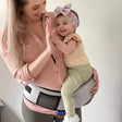 Mother wearing cream baby hip carrier while giving high-five to toddler.