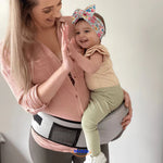 Mother wearing cream baby hip carrier while giving high-five to toddler.