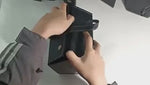 Demo of a black foldable multi-compartment eyeglasses case being held by its handle, showing portability.