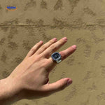 Silver ring watch with black square dial on hand model.